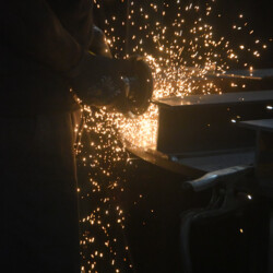 A person uses an angle grinder to cut a metal beam, producing a shower of sparks.