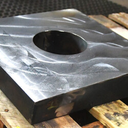 A square metal plate with a circular hole in the center, resting on a wooden pallet.