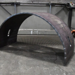 A curved metal plate with slots on a shop floor in front of a metal press.