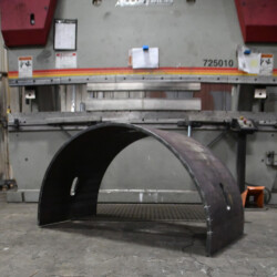 A curved metal sheet with two slots sits on the floor in front of a large industrial press brake.