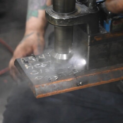 A metal drill press creates smoke while boring a hole into a piece of steel.